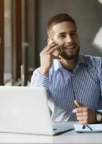 Scientific engineer smiling on the phone, with a notebook, pens, and a laptop in front of him